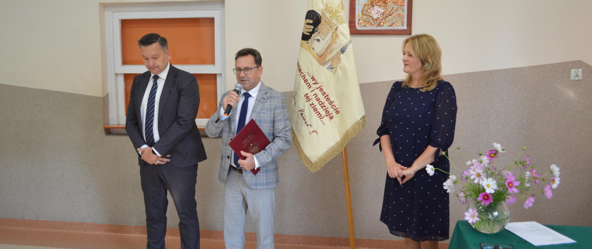 Na zdjęciu starosta i wicestarosta przemawiają do społeczności szkolnej w obecności dyrektora Zespołu Szkół Specjalnych im. Jana Pawła II w Sokołowie Podlaskim. Tuż obok ustawiony na drewnianym statywie sztandar szkoły.