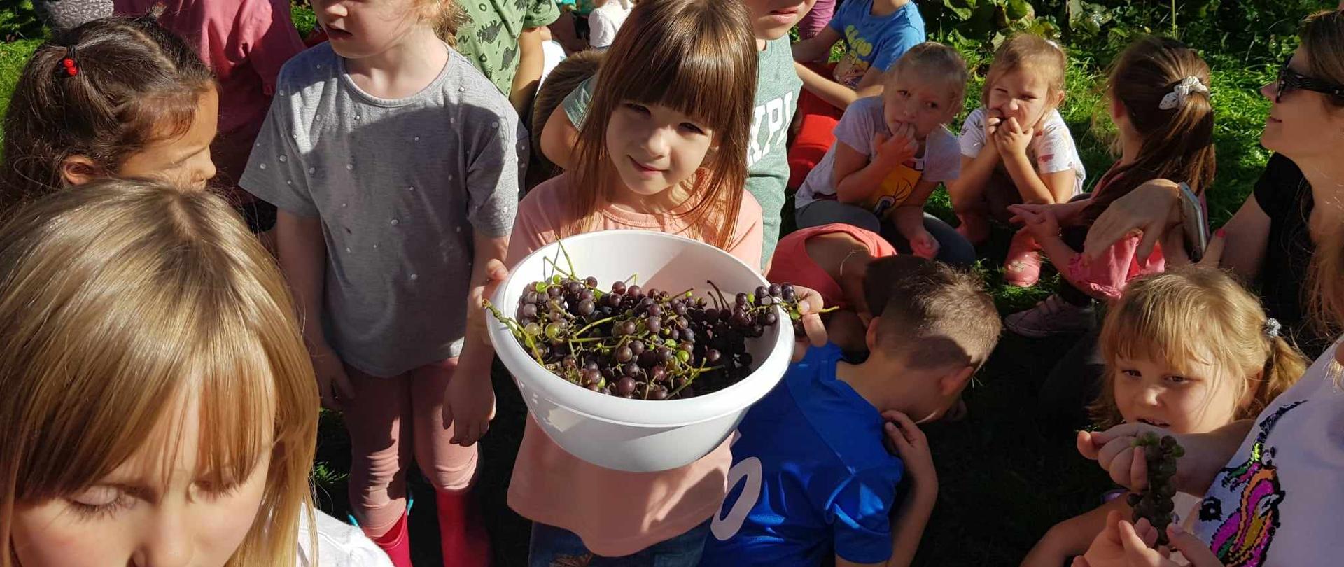 W centrum grupy dzieci stoi dziewczynka trzymająca miskę wypełnioną winogronami.