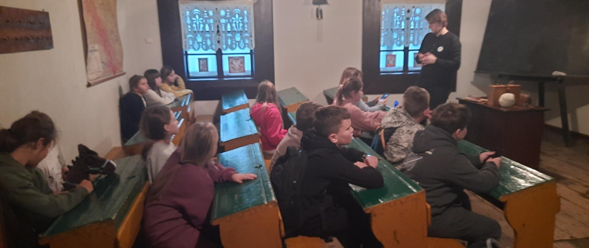 Uczniowie w Muzeum Etnograficznym w Krakowie siedzą w ławkach w pomieszczeniu zorganizowanym na wzór bardzo starej sali lekcyjnej. Przed nimi stoi pracownik muzeum i opowiada im o czymś.