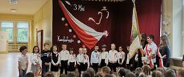 Przedszkolaki ubrani na galowo czyli białe koszule, granatowe spodnie lub spódniczki występują w akademii, na koszulkach mają przypięte biało-czerwone kotyliony. Stoją na tle bordowej zasłony, na której przypięte są biało-czerwone symbole Polski czyli flaga, orzeł w złotej koronie, kwiaty oraz napisy "3 maj" i "Konstytucja". Po prawej stronie stoi poczet sztandarowy ze sztandarem szkoły, przed występującymi widać siedzących uczniów jako widzów