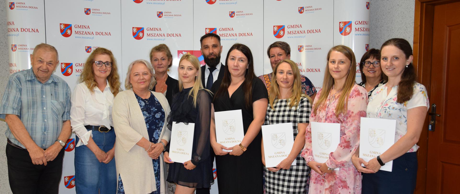 Wójt Gminy Mszana Dolna z nauczycielkami, które w drugim dniu postępowania otrzymały awans zawodowy na stopień nauczyciela mianowanego wraz z komisją egzaminacyjną, w skład której wchodzili: dyrektor szkoły, wizytator z kuratorium oświaty oraz dwóch ekspertów z danych przedmiotów. Stoją na tle ścianki z herbem Gminy Mszana Dolna.