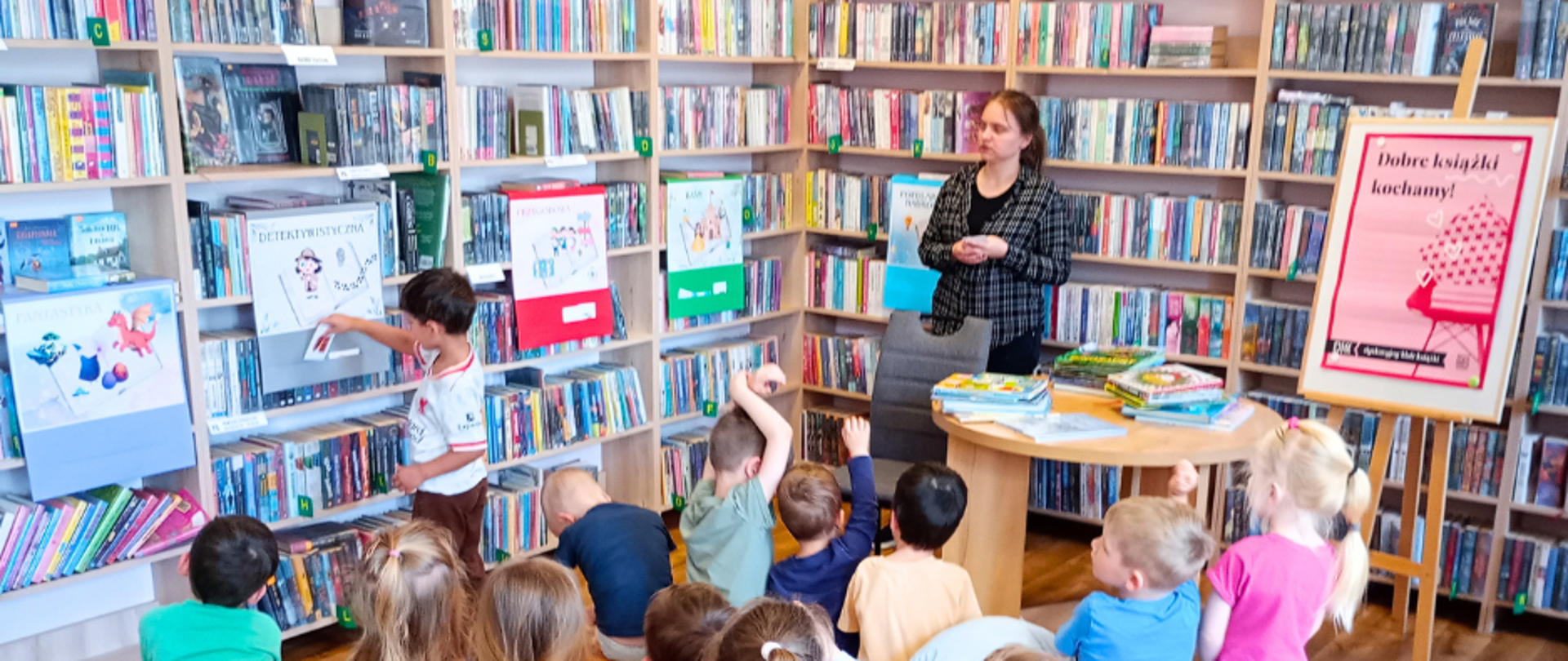 Na zdjęciu dzieci siedzą na ławeczkach w bibliotece i zgłaszają się do udziały w zabawie polegającej na przyporządkowywaniu elementów narysowanych na karteczkach pokazywanych przez prowadzącą do odpowiednich plakatów z gatunkami literackimi.