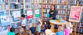 Na zdjęciu dzieci siedzą na ławeczkach w bibliotece i zgłaszają się do udziały w zabawie polegającej na przyporządkowywaniu elementów narysowanych na karteczkach pokazywanych przez prowadzącą do odpowiednich plakatów z gatunkami literackimi.