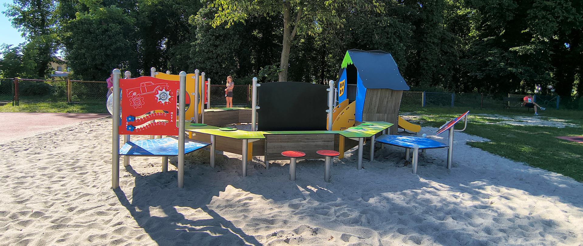 Szerokie ujęcie piaszczystego placu zabaw usytuowanego w cieniu dużego drzewa. W tle widoczny jest metalowy płot oraz dojrzały park z gęstą roślinnością.