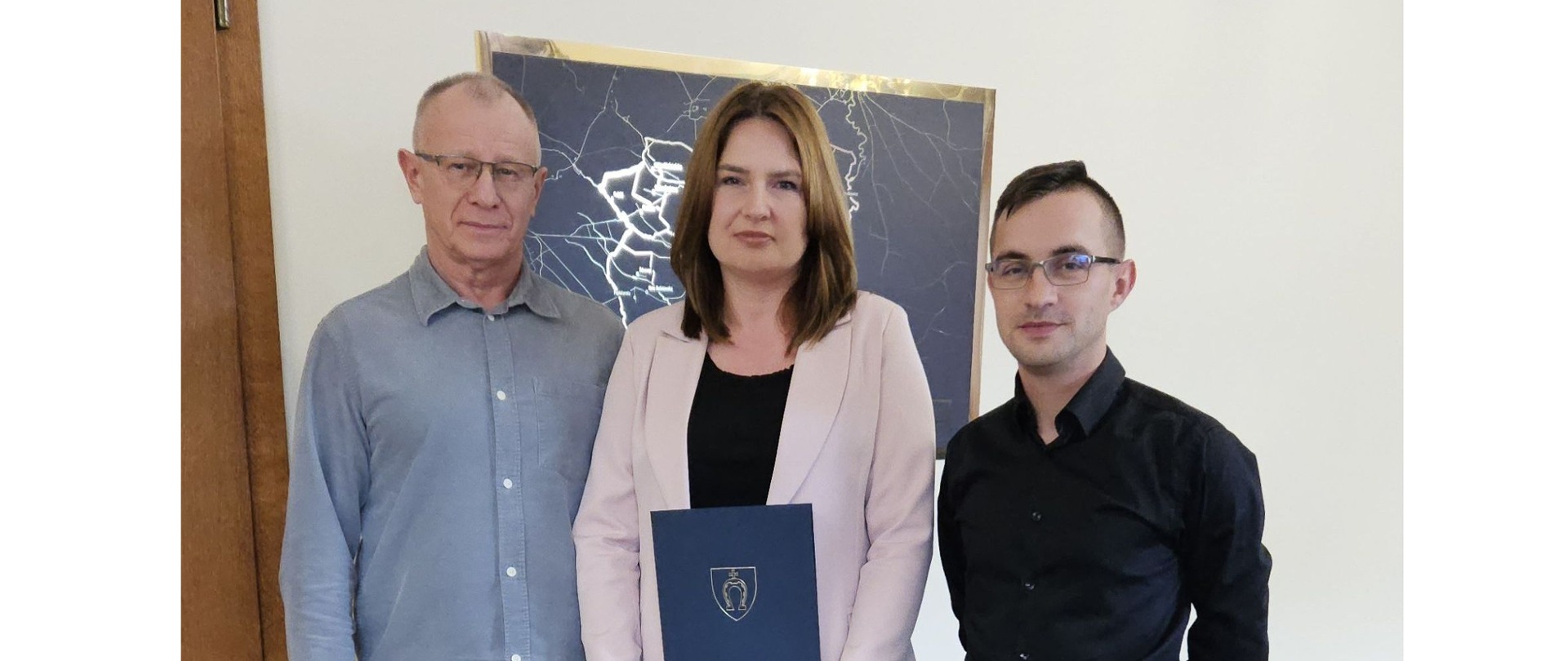 W gabinecie na tle jasnej ściany stoi dwóch mężczyzn i pośrodku kobieta trzymająca w ręku ozdobną niebieską teczkę z herbem. Mężczyzna po lewej ubrany w niebieską koszulę z białymi guzikami, po prawej w czarną koszulę a kobieta w różowy żakiet. Na ścianie wisi czarny obraz w złotej ramce.