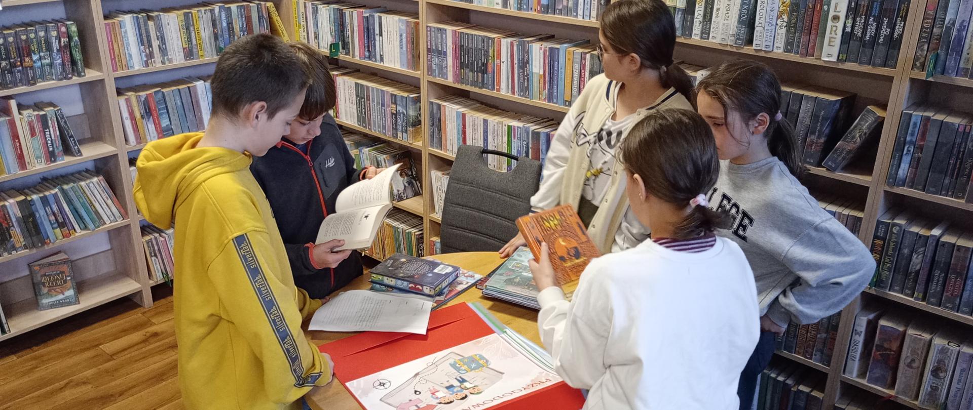 Na zdjęciu widać dzieci stojące dookoła stołu, na którym rozłożone są książki i plakaty poświęcone różnym gatunkom literackim. Dzieci przeglądają te materiały.