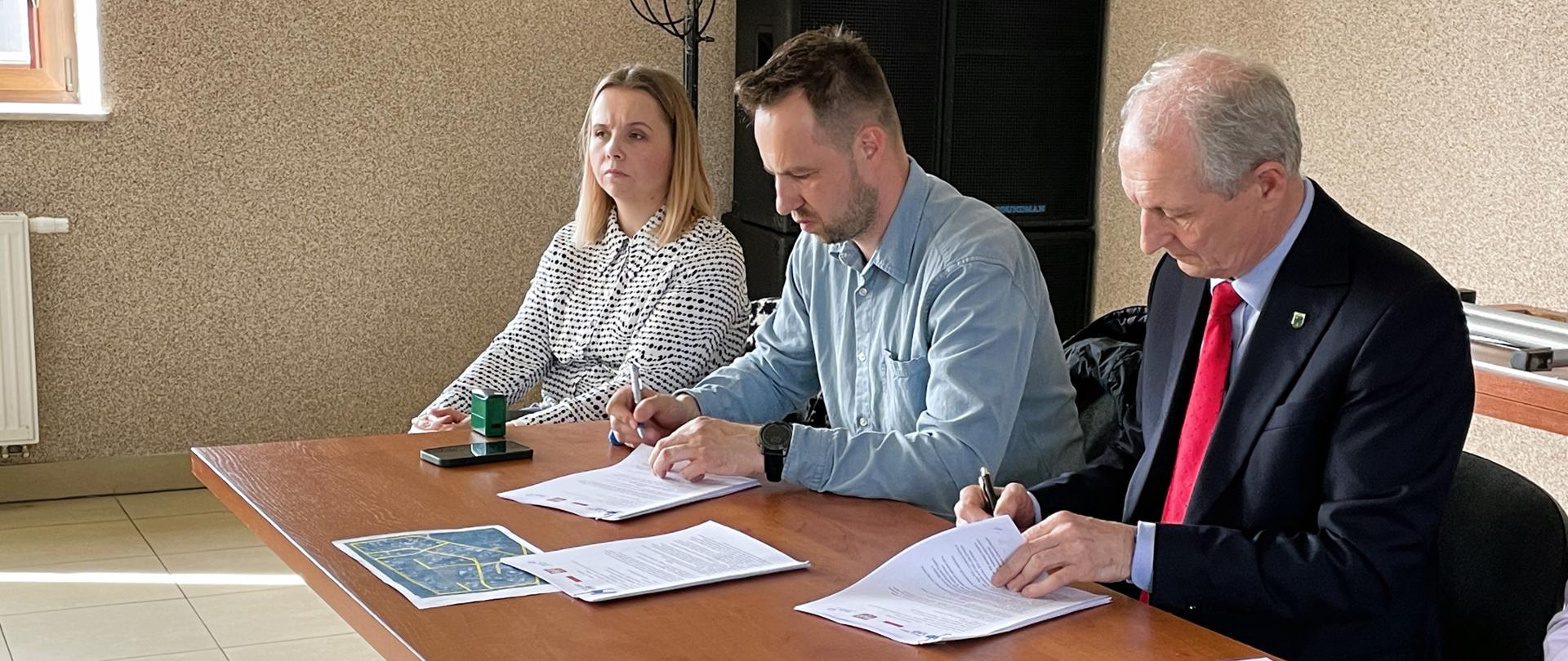 Dwóch mężczyzn podpisujących umowę, obok siedzi kobieta i mężczyzna.