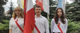 Na zdjęciu znajduje się poczet sztandarowy szkoły. W środku stoi chłopak trzymający sztandar. Ubrany jest w białą koszulę i czarne spodnie. Między nim stoją dwie dziewczyny. Obie ubrane są w białe koszule i czarne spódnice.