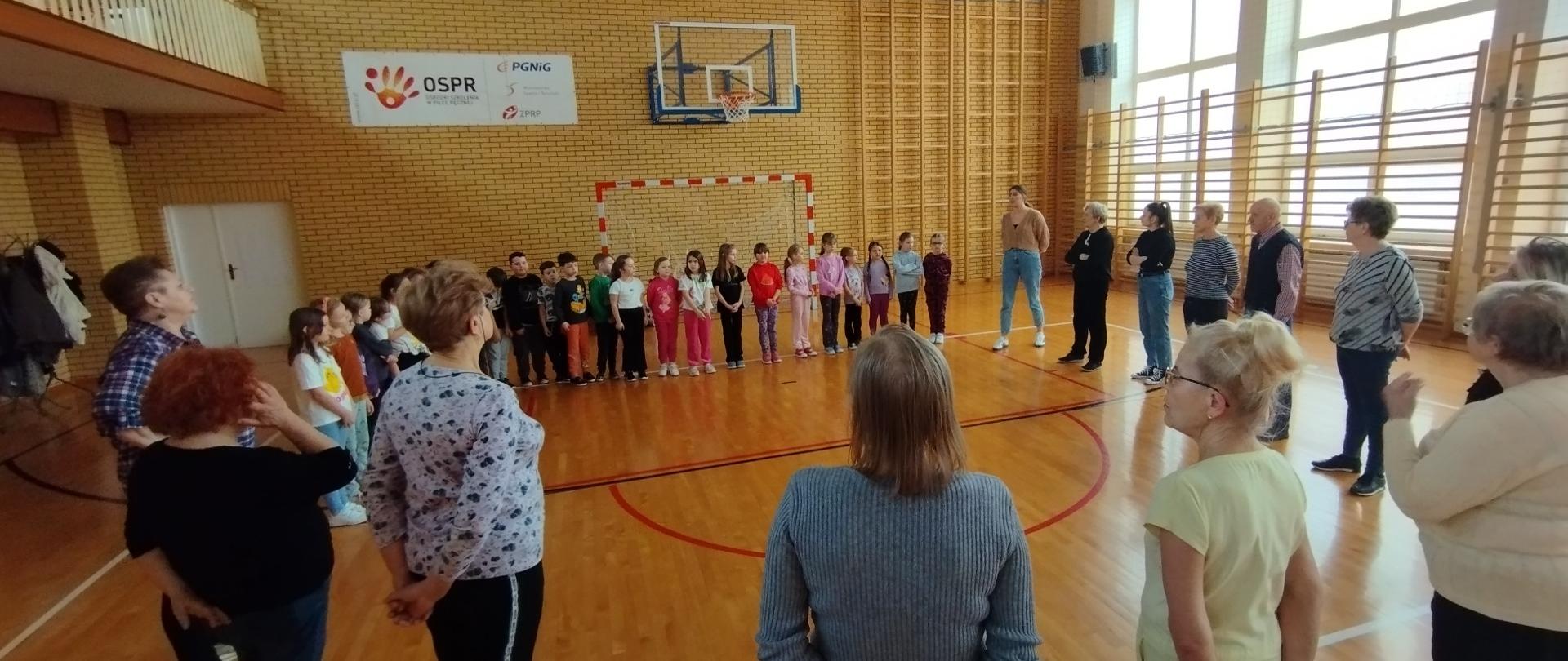 Członkowie Klubu Senior+ wraz z uczniami Szkoły podstawowej na sali gimnastycznej podczas spotkania integracyjnego
