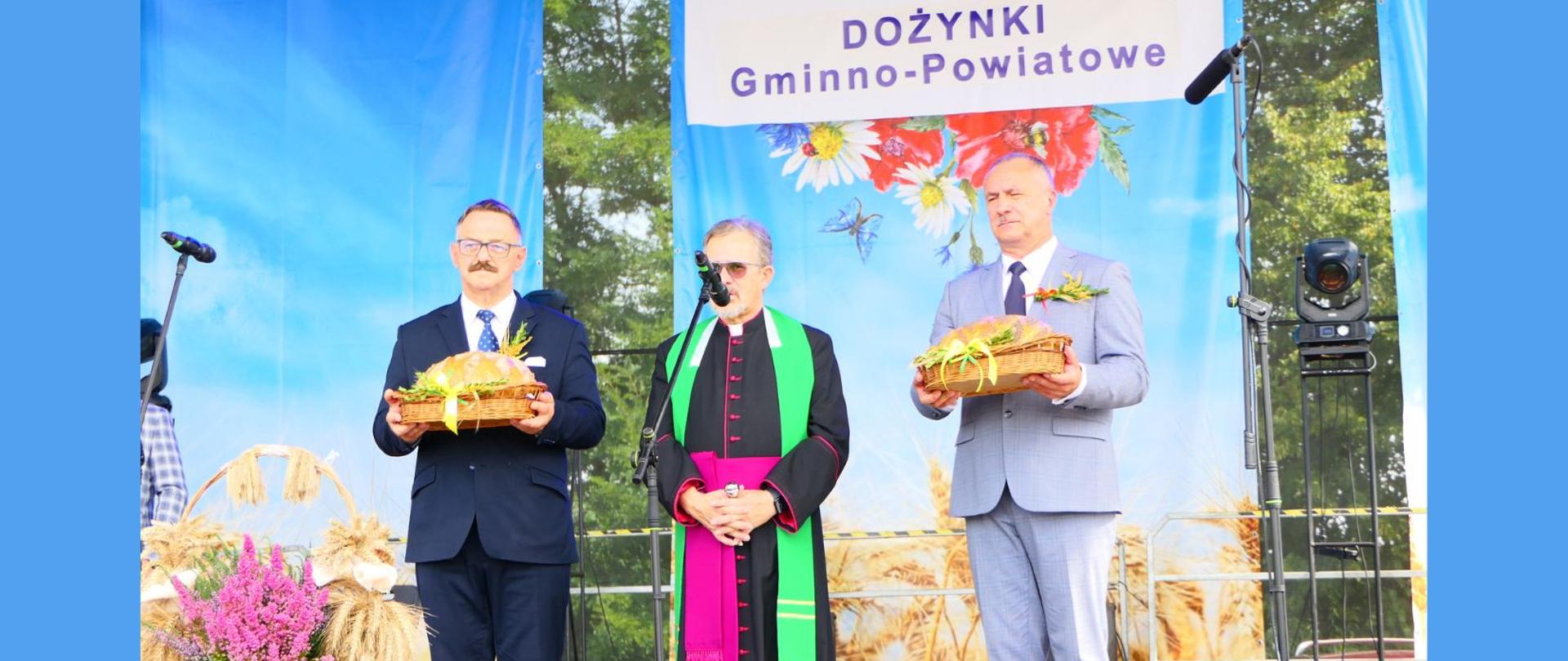 Starosta Lipski oraz Wójt Sienna trzymają chleby dożynkowe. Między nimi stoi proboszcz parafii Sienno Andrzej Cieszkowski. W tle błękitny baner wydarzenia z napisem Dożynki Gminno-Powiatowe i przewodnim motywem graficznym z chabrami, rumiankami i makami.