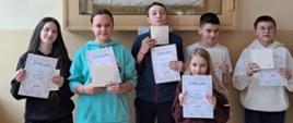Trzy uczennice i trzech uczniów będących zwycięzcami w konkursie stoi pod gablotką ze sztandarem szkoły przedstawiającym Świętą Kingę, trzymają w rękach dyplomy