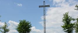 Metalowy krzyż, przed krzyżem głaz, obok krzyża młode drzewka