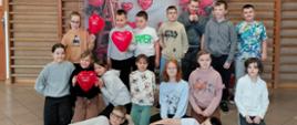 Uczniowie klasy trzeciej pozują do wspólnego zdjęcia na specjalnie przygotowanej ściance walentynkowej. Za nimi, na drabinkach wiszą czerwone balony w kształcie serc.