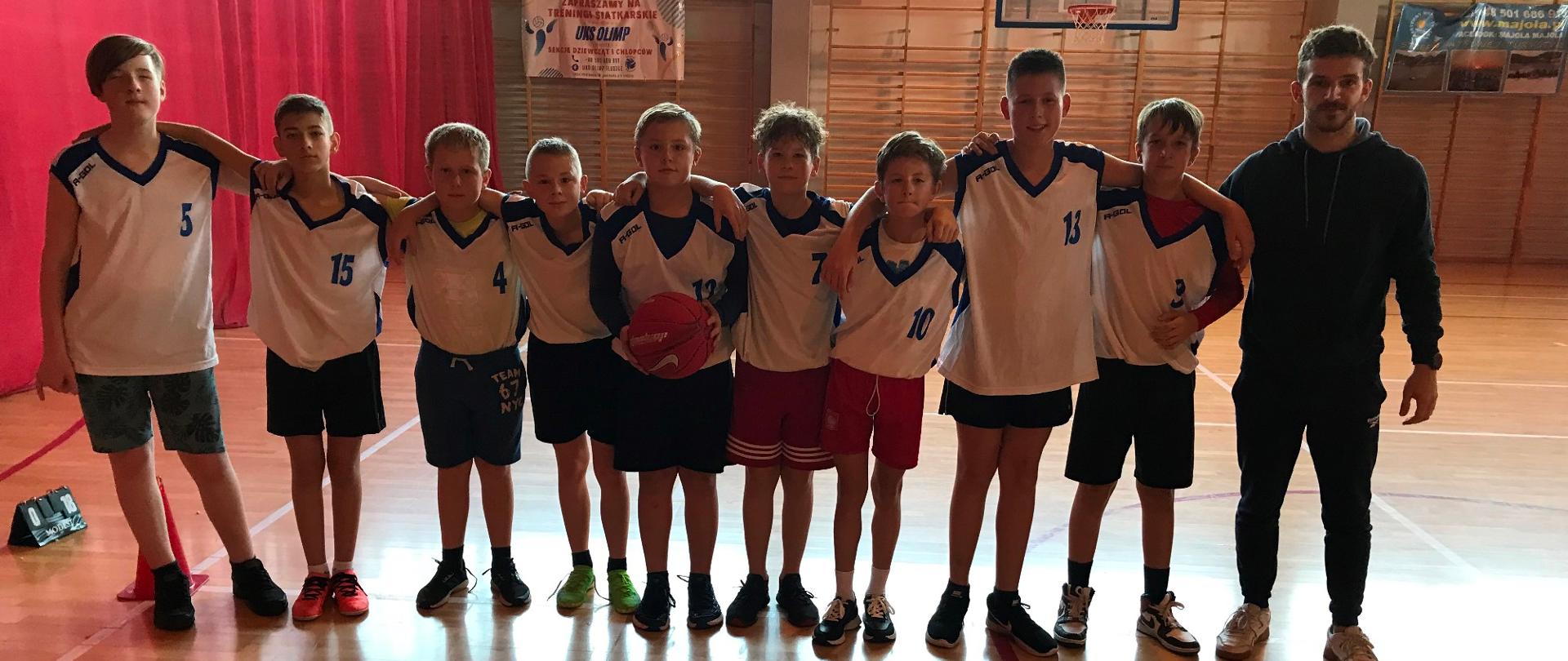 Chłopcy w białych koszulkach i spodenkach stoją obok siebie i obejmują się za ramiona. Obok stoi mężczyzna w stroju sportowym