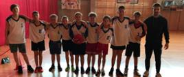 Chłopcy w białych koszulkach i spodenkach stoją obok siebie i obejmują się za ramiona. Obok stoi mężczyzna w stroju sportowym