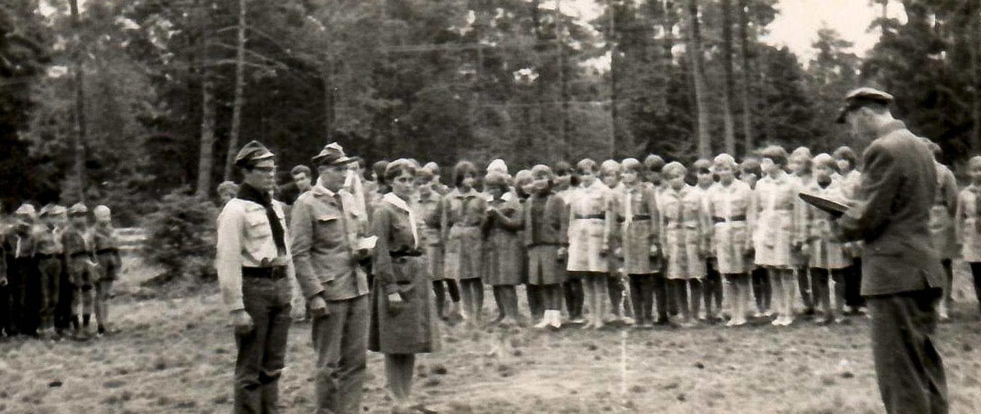 Czarno-biała fotografia grupowa harcerzy z Konstantynowa Łódzkiego sprzed 1975 roku. Grupa stoi w równych rzędach w mundurach harcerskich. W tle las.