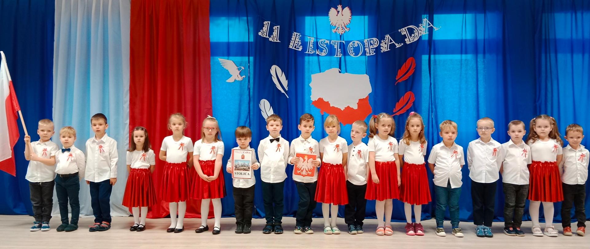 Dzieci ubrane na galowo (dziewczynki - białe koszulki i czerwone spódniczki, chłopcy - białe koszule i czarne spodnie) na tle dekoracji z napisem 11 listopada 
