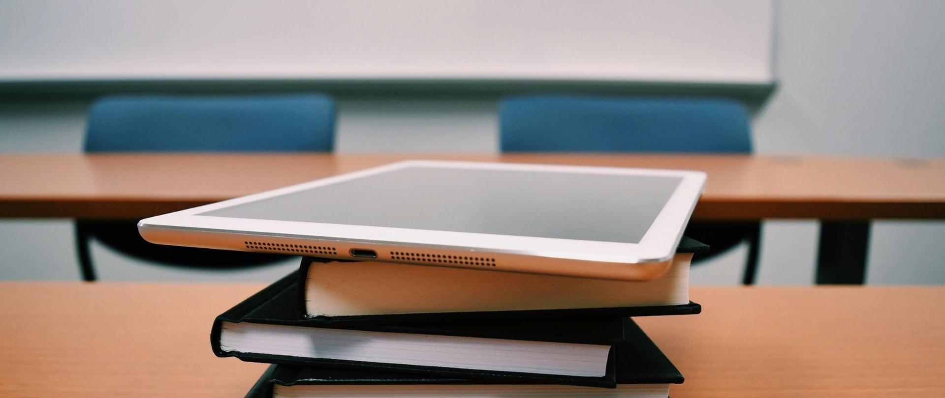 Zdjęcie przedstawia salę lekcyjną. W tle tablica, a na pierwszym planie biurko a na nim kilka książek położonych jedna na drugiej a na niuch tablet.