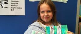 Dziewczynka z klasy 5 trzyma w jednej ręce wypożyczoną z biblioteki książkę przedstawiającą 2 pary sznurowanych butów na zielonym tle, w drugiej ręce wykonany przez siebie obrazek z takim samym motywem