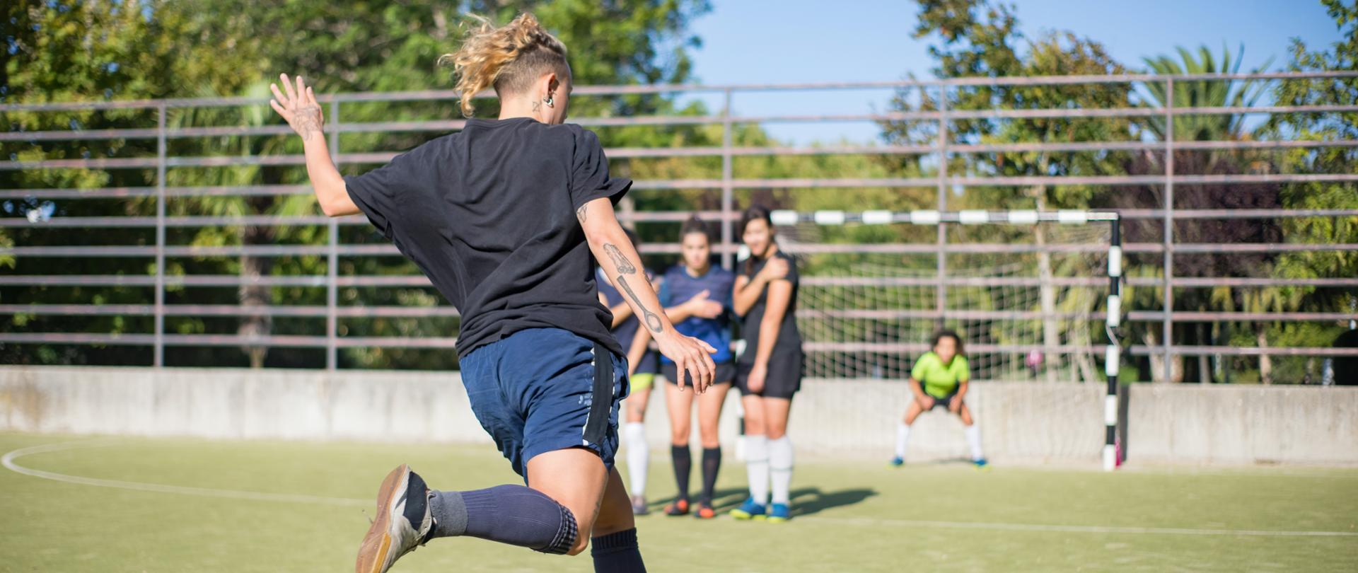 Sporty young woman kicking ball on summer day. Sportswoman in dark uniform kicking ball in direction of football goals, teammates in background. Sport, leisure, active lifestyle concept