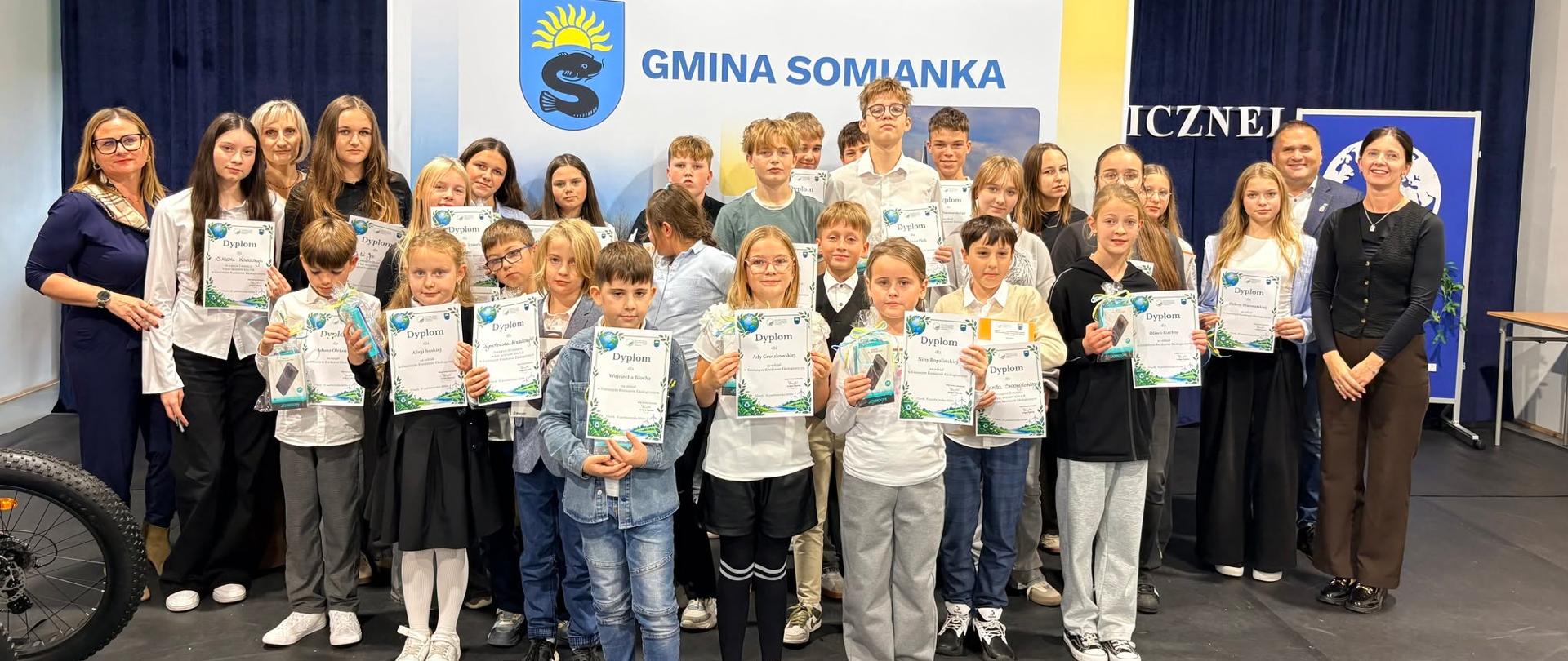 Zdjęcie pokazuje wszystkich uczestników konkursu o wiedzy ekologicznej . Dzieci stoją uśmiechnięci, w rękach trzymają dyplomy za udział w konkursie . W tle baner gminy Somianka. 