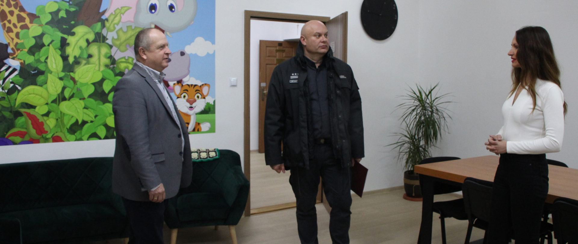 Zdjęcie przedstawia wnętrze pomieszczenia PCPR. Na fotografii widać trzy osoby: od lewej Wicestarostę Powiatu Zambrowskiego, Dyrektora Zakładu Karnego w Czerwonym Borze oraz Starostę Powiatu Zambrowskiego. W tle znajduje się kolorowa fototapeta z motywem zwierzęcym, ciemnozielona kanapa oraz dwa fotele w takim samym kolorze. Po prawej stronie stoi stół z krzesłami, a na podłodze roślina doniczkowa.