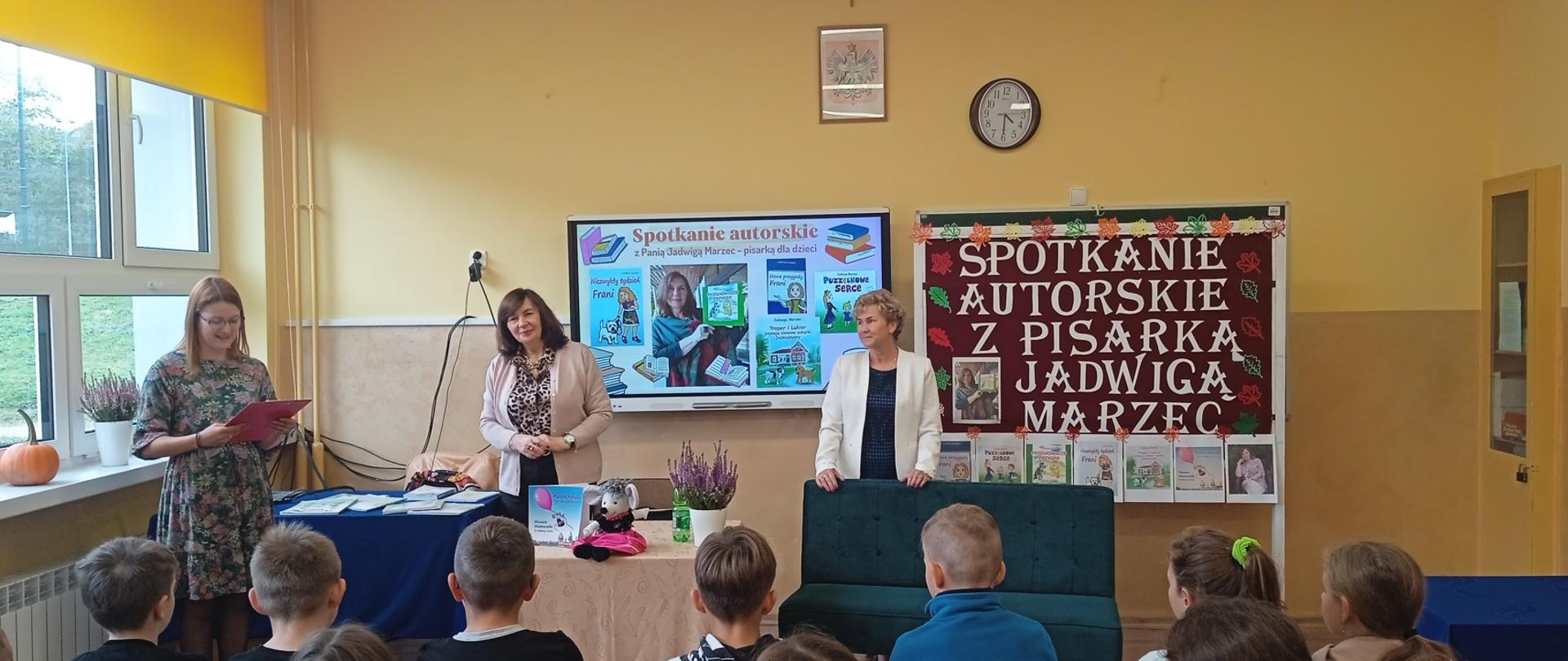 Spotkanie autorskie z pisarką Panią Jadwigą Marzec - na środku klasy stoi pisarka, po lewej stronie kierownik Gminnej Biblioteki Publicznej Pani Dominika Plata, po prawej - bibliotekarka ze Szkoły w Nowej Wsi Pani Krystyna Borek, w tle monitor z prezentacją i gazetka promujące pisarkę, na przodzie uczniowie klasy czwartej
