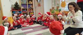 Dzieci ubrane na czerwono, w czapkach świętego Mikołaja siedzą na dywanie w kółku, obok nich pani nauczycielka