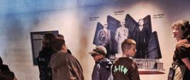 Dzieci i kobieta stoją w pomieszczeniu w którym na ścianie nadrukowane są 3 postaci mężczyzn o imieninach Karol, Emil i Jan Wedel