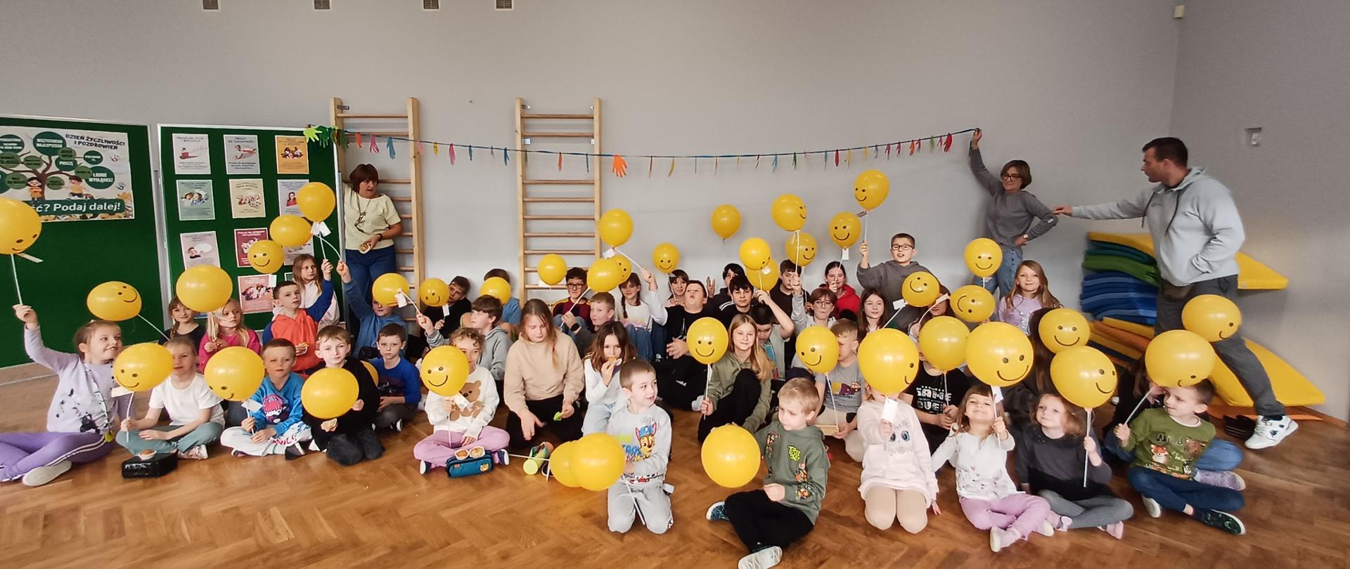 Zdjęcie przedstawia uczniów szkoły podczas obchodów Dnia Życzliwości i Pozdrowień trzymających balony z emotikoną uśmiechu. Na dalszym planie nauczyciele trzymający łańcuch z wyciętych kolorowych łapek.