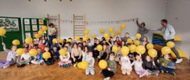 Zdjęcie przedstawia uczniów szkoły podczas obchodów Dnia Życzliwości i Pozdrowień trzymających balony z emotikoną uśmiechu. Na dalszym planie znajdują się nauczyciele trzymający łańcuch z wyciętych kolorowych łapek.