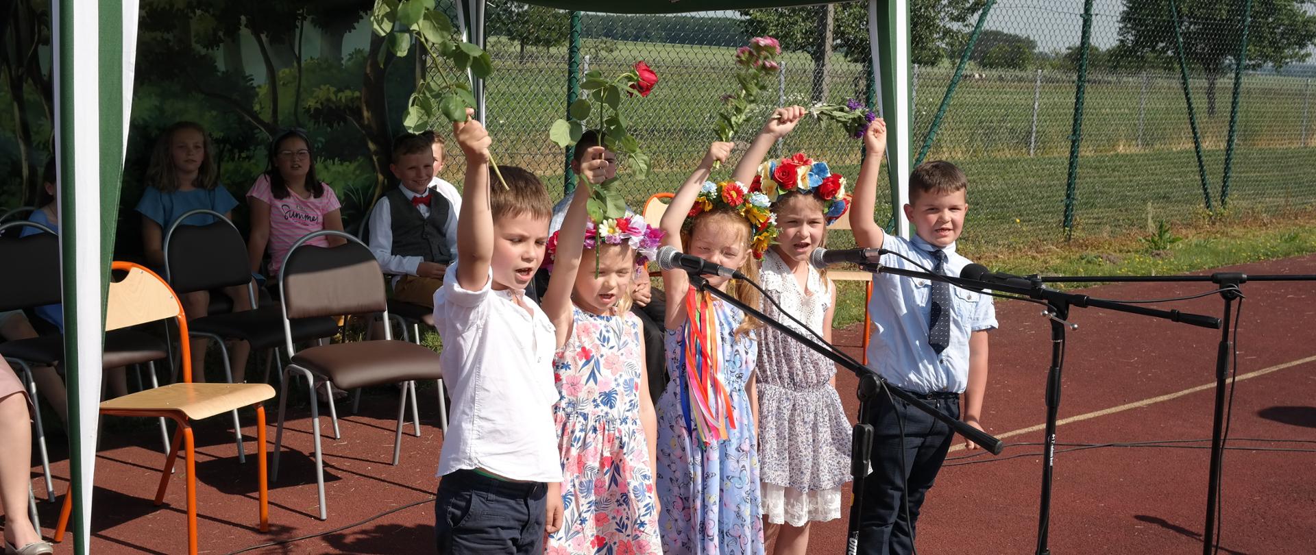 Najmłodsza grupa wokalna w składzie pięcioosobowym, w plenerze, wykonuje piosenkę pt. "Idzie wiosna. dzieci kolorowo ubrane z wiankami na głowie.