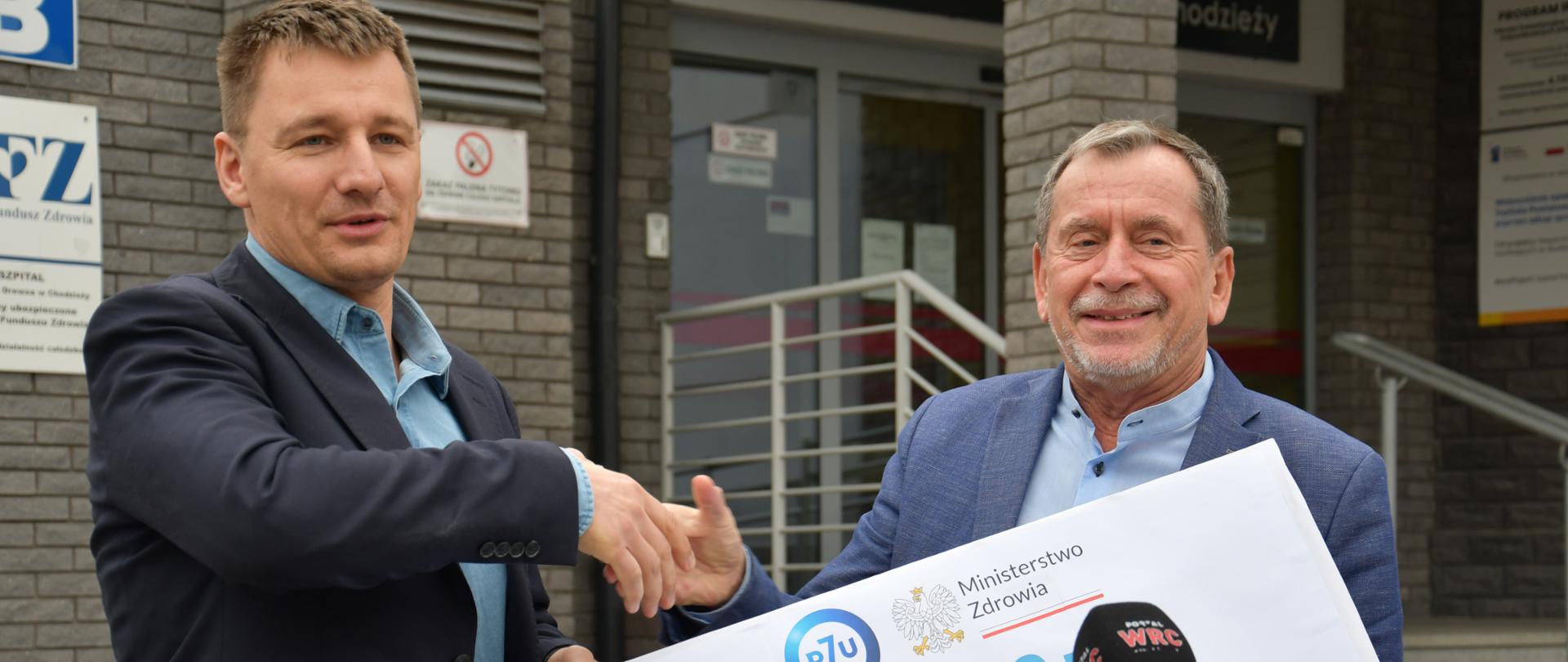 2 osoby w garniturach stojące przed wejściem do budynku z symbolicznym czekiem na 2 000 000 zł
