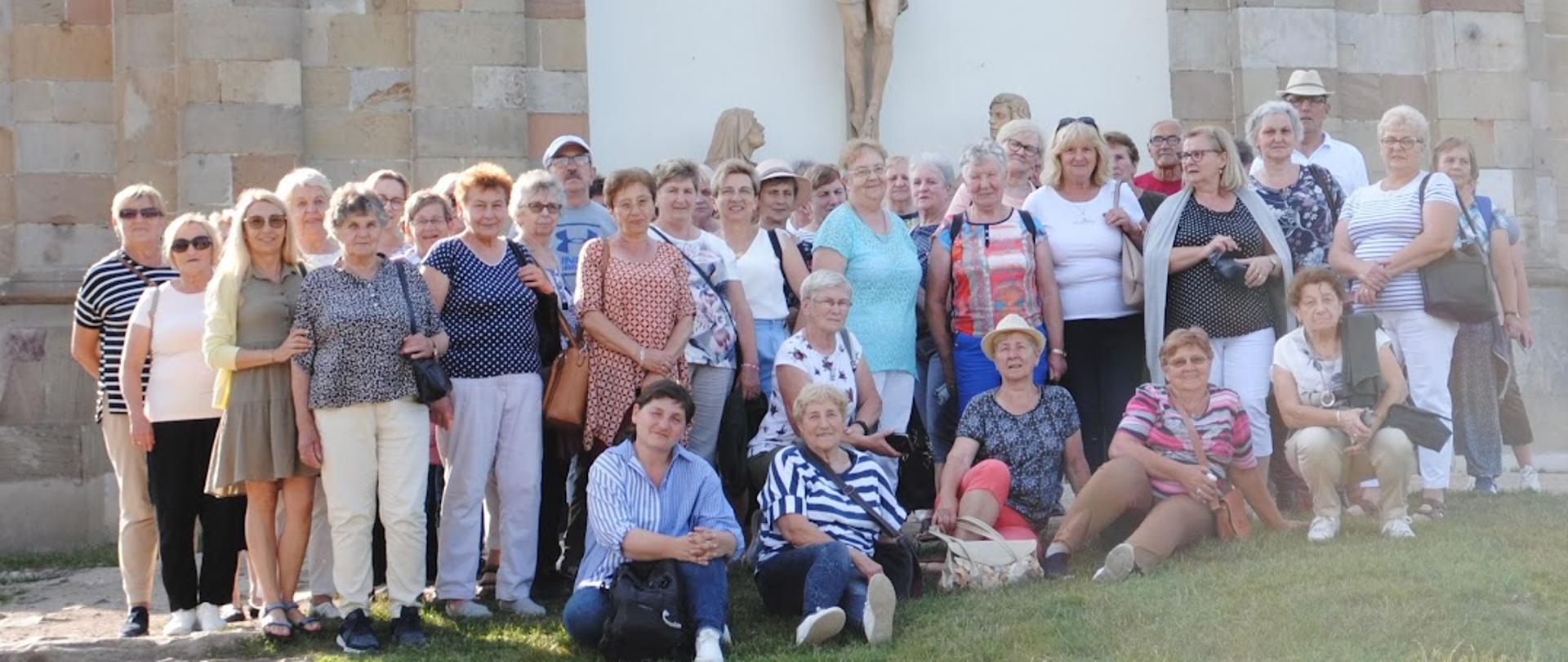 mężczyźni i kobiety pozują do zdjęcia stoją i kucają pod ściana jakiejś budowli na ścianie duży krzyż 