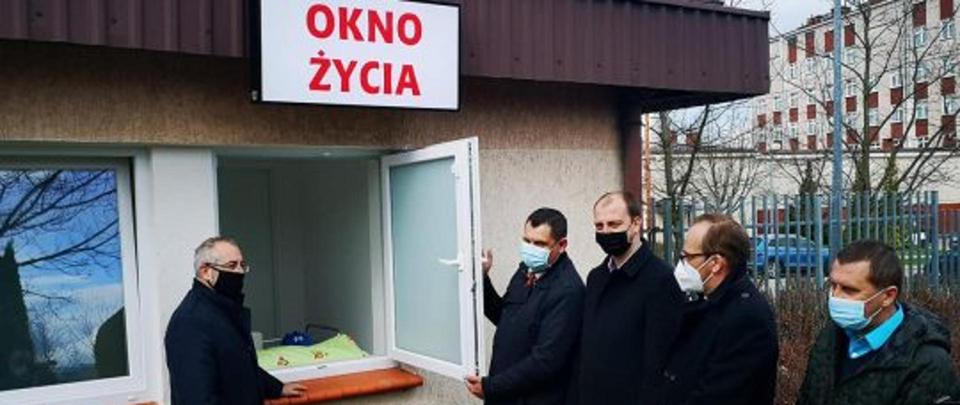 Zdjęcie przedstawia chwilę tuż po uroczystym otwarciu Okna Życia w powiecie przasnyskim. Starosta Krzysztof Bieńkowski (pierwszy od lewej) prezentuje zebranym gościom oraz mediom funkcjonalność Okna Życia. Na zdjęciu kolejno po staroście również: Dyrektor przasnyskiego Szpitala Grzegorz Magnuszewski, Burmistrz Przasnysza Łukasz Chrostowski, Przewodniczący Rady Powiatu Przasnyskiego Paweł Szczepkowski, Pełnomocnik Powiatu w zakresie Samorządowej Karty Praw Rodzin Zbigniew Makowski. Okno Życia zostało usytuowane w portierni przy wjeździe na teren Szpitala w Przasnyszu – od strony Urzędu Skarbowego.