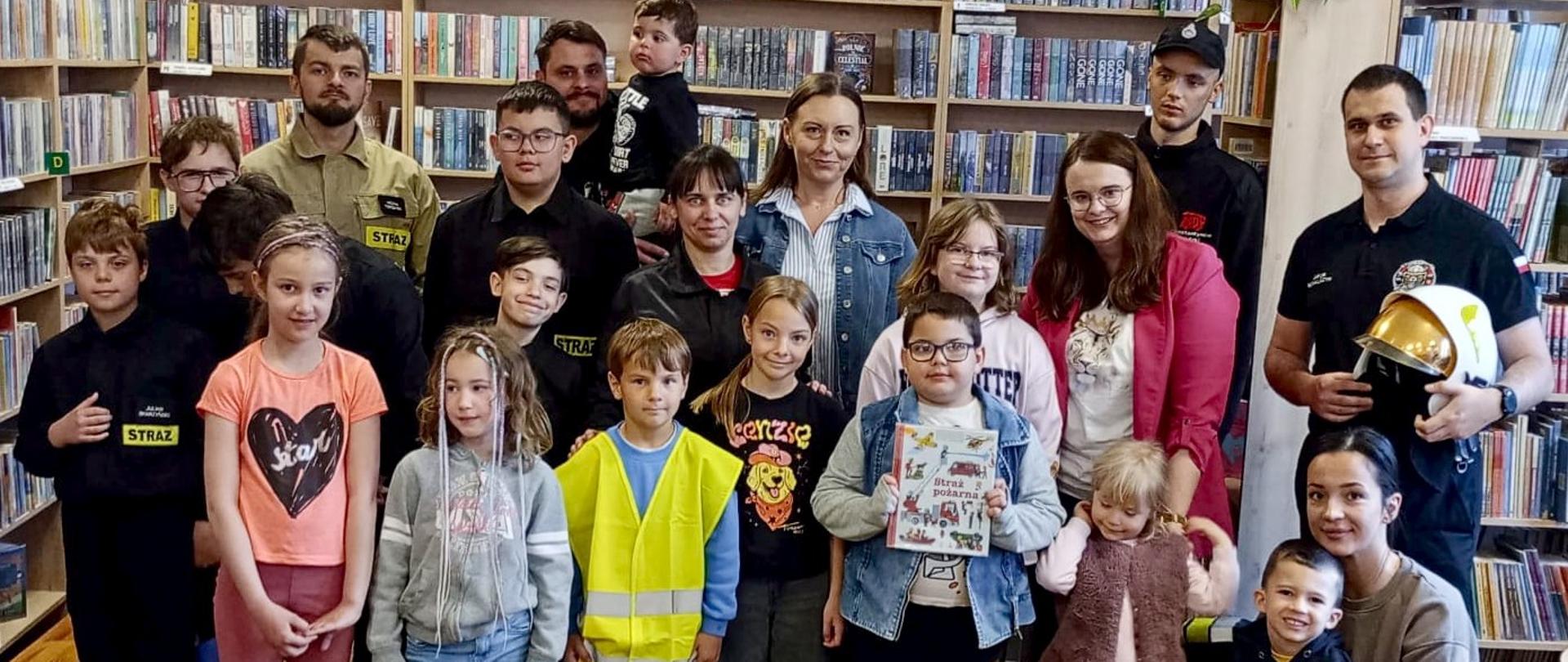 Na zdjęciu dzieci pozują ze strażakami z OSP Konstantynów na tle bibliotecznych regałów.