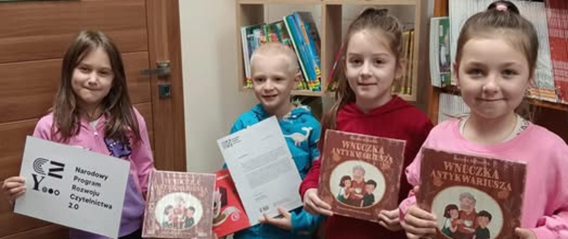 W bibliotece przed regałem z nowościami stoją uczniowie klasy 1, 3 dziewczynki i 1 chłopiec, w ręku trzymają książki otrzymane w ramach kampanii, jedna dziewczynka trzyma logo Narodowego Programu Rozwoju Czytelnictwa