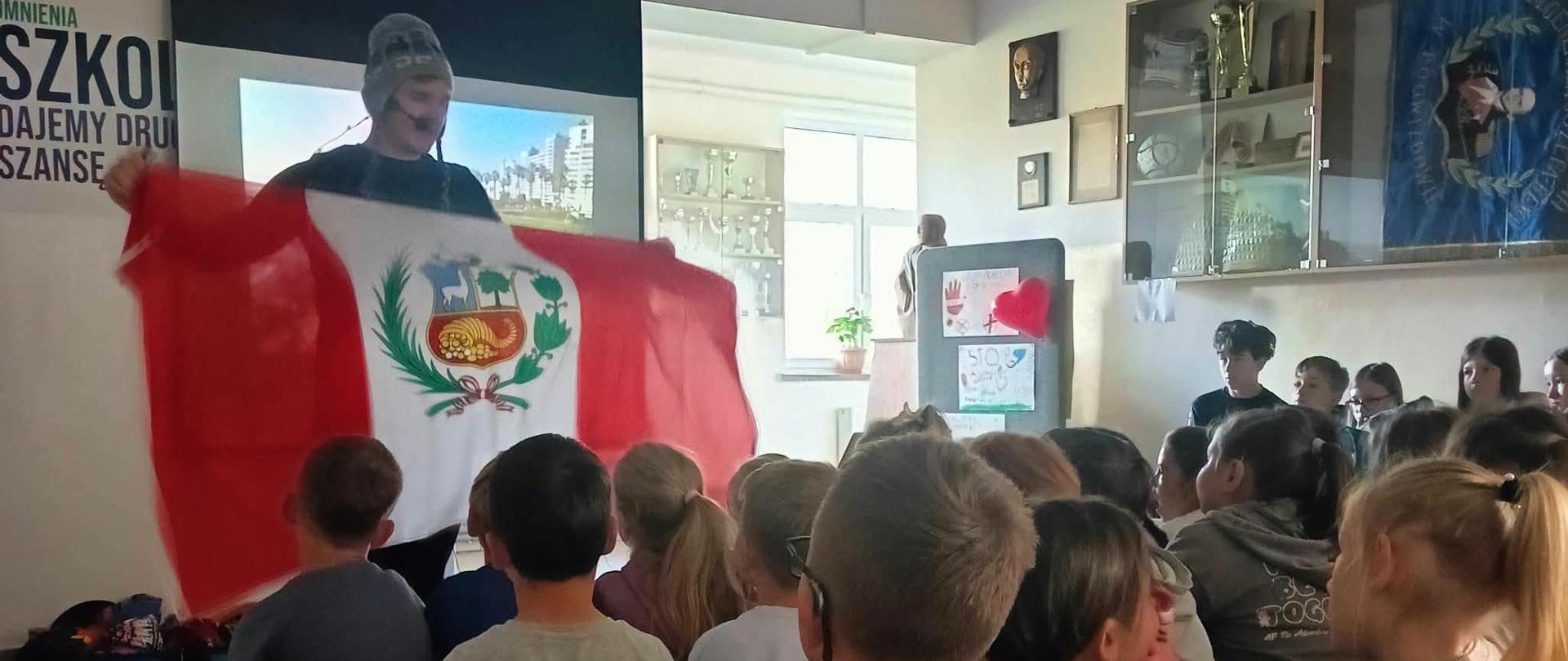Na zdjęciu widać spotkanie z dziećmi w sali szkolnej. Na pierwszym planie siedzi grupa uczniów, którzy z zainteresowaniem oglądają prezentację. Przed nimi stoi mężczyzna w czapce, trzymający dużą flagę Peru w charakterystycznych czerwono-biało-czerwonych barwach, z godłem pośrodku.
W tle znajduje się ekran, na którym wyświetlana jest prezentacja, a także gabloty z pucharami i trofeami. Na ścianach wiszą dekoracje oraz pamiątki. W pomieszczeniu panuje przyjazna atmosfera, a dzieci uważnie obserwują prowadzącego.