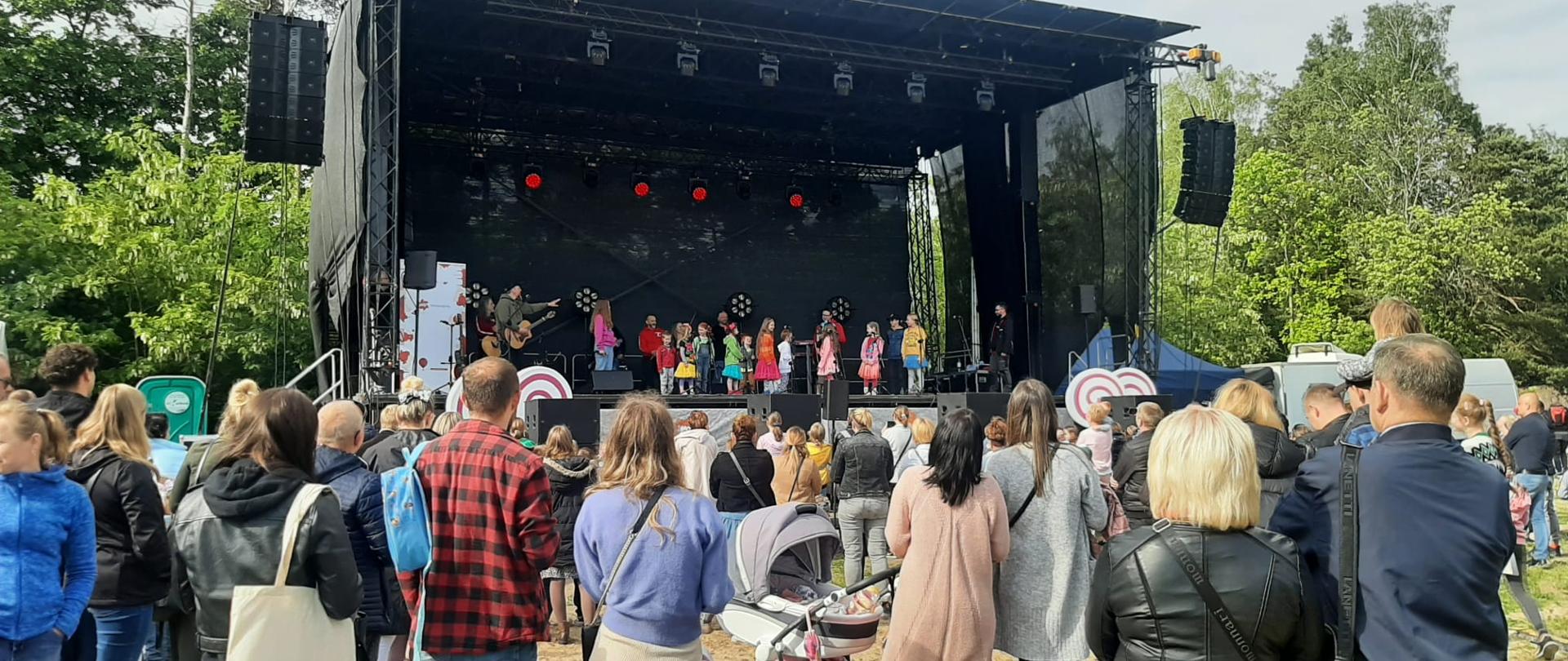 Scena na tle drzew, pod sceną tłum ludzi, na scenie zespół Arka Noego składający się z ponad 20 dzieci i zespół muzyków.