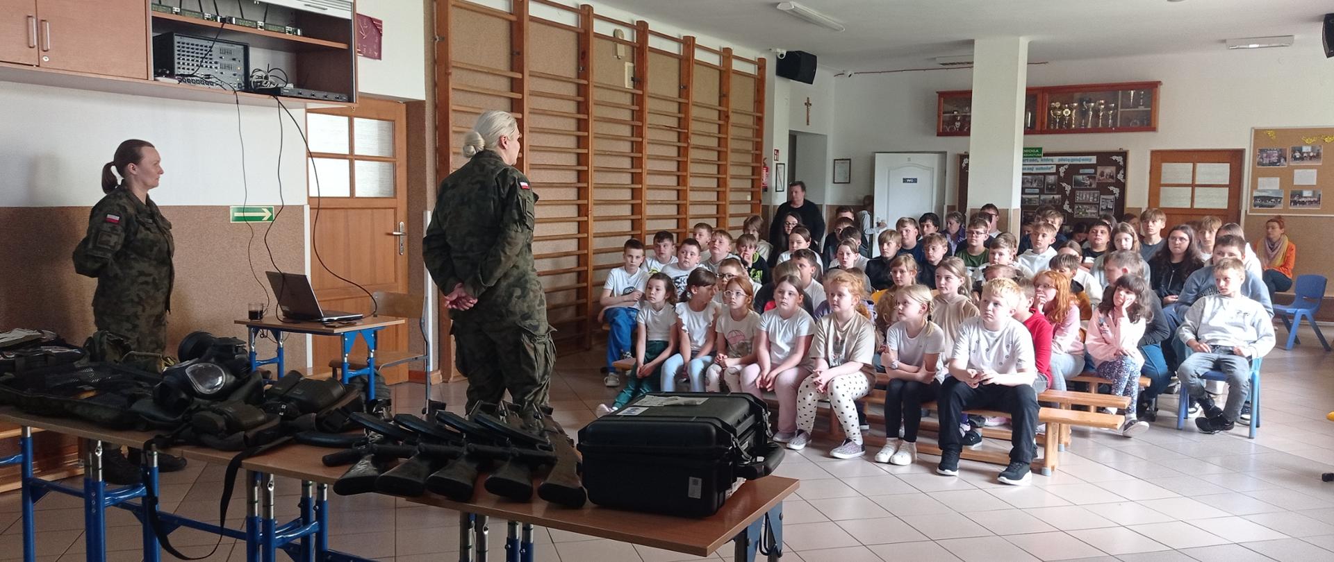 Na korytarzu szkolnym siedzą uczniowie klas starszych. Ich twarze są skierowane ku dwóm kobietom ubranym w mundury wojskowe.