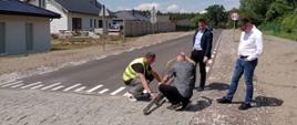droga z progiem zwalniającym z kostki brukowej, na drodze kucają 2 osoby, jedna stoi, kolejna osoba stoi na poboczu, po prawej stronie drogi gęste drzewa, po lewej stronie drogi budynki i płot betonowy
