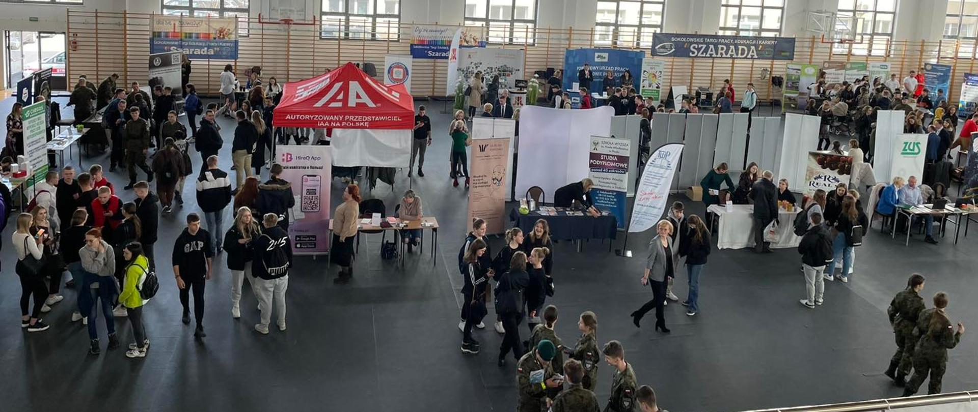 Widok z góry na halę sportową Zespołu Szkół Nr 1. Na fotografii widać poustawiane na całym obszarze hali stoiska promocyjne, a pomiędzy nimi młodzież oraz uczestników Targów.