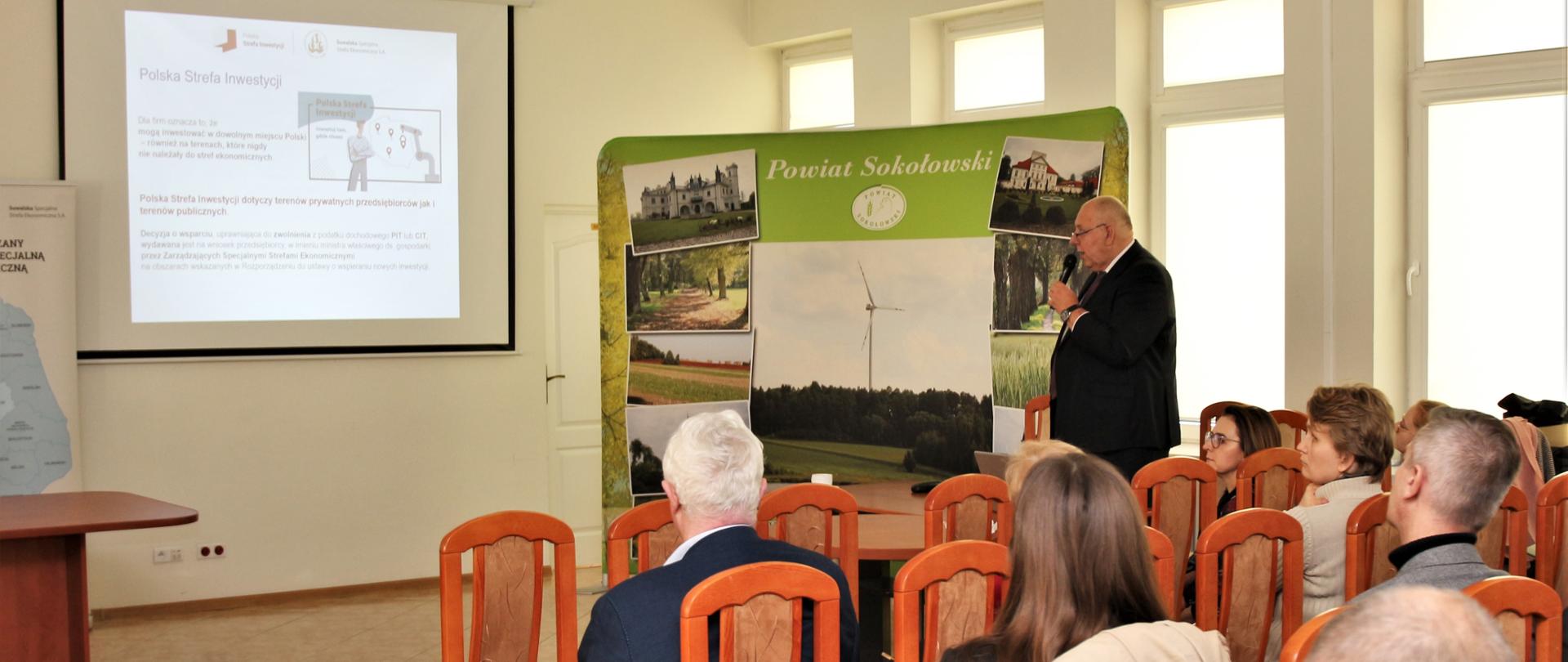 Na fotografii jest przedstawiony Prezes Zarządu SSSE S. A. Leszek Dec. który wyjaśnia czym jest Polska Strefa Inwestycji. W tle widać ściankę Powiatu Sokołowskiego oraz slajd wyświetlany przez projektor. Po prawej stronie zgromadzeni ludzie.