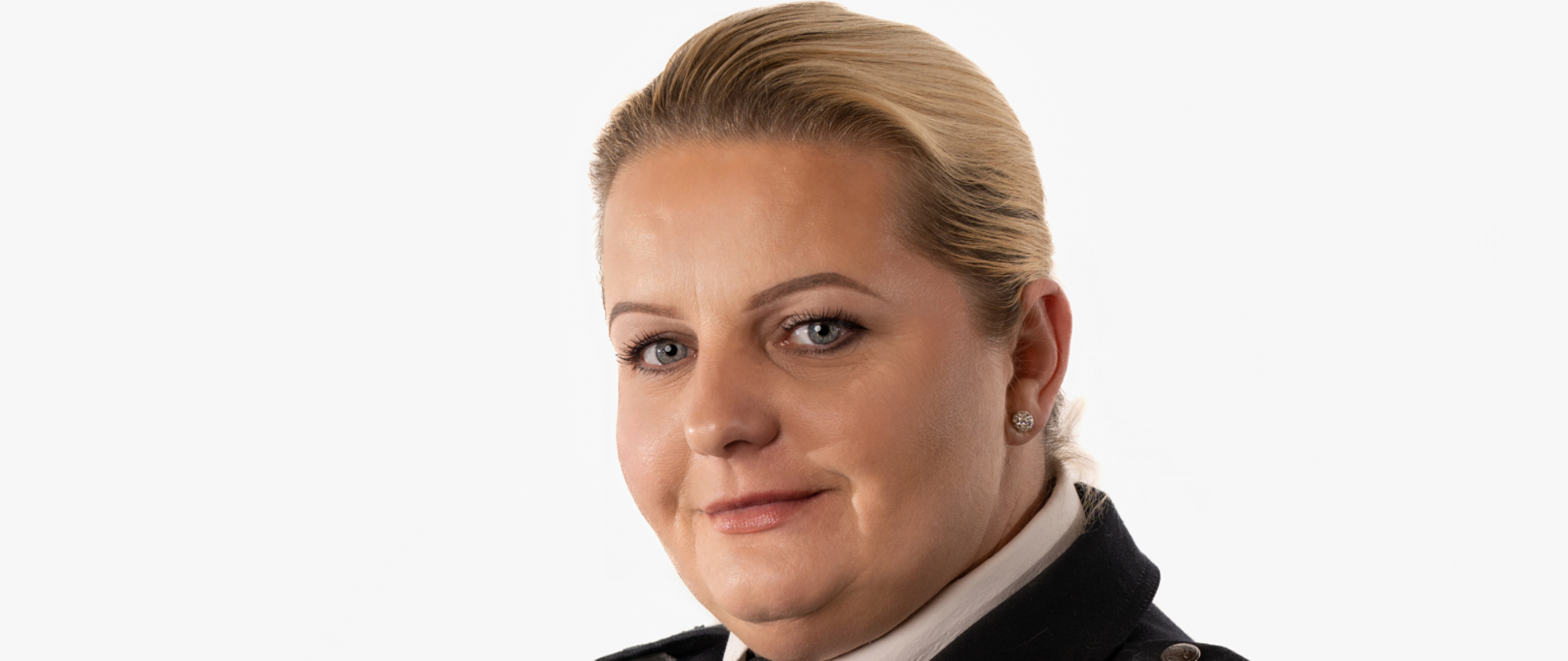 Blondynka o niebieskich oczach, włosy spięte z tyłu, na twarzy lekki uśmiech. Ubrana w strażacki mundur galowy, do tego biała koszula i krawat