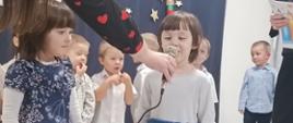 Kącik artystyczny w wykonaniu przeszłych przedszkolaków - przy głosie dziewczynka, recytuje kwestię stojąc przed mikrofonem