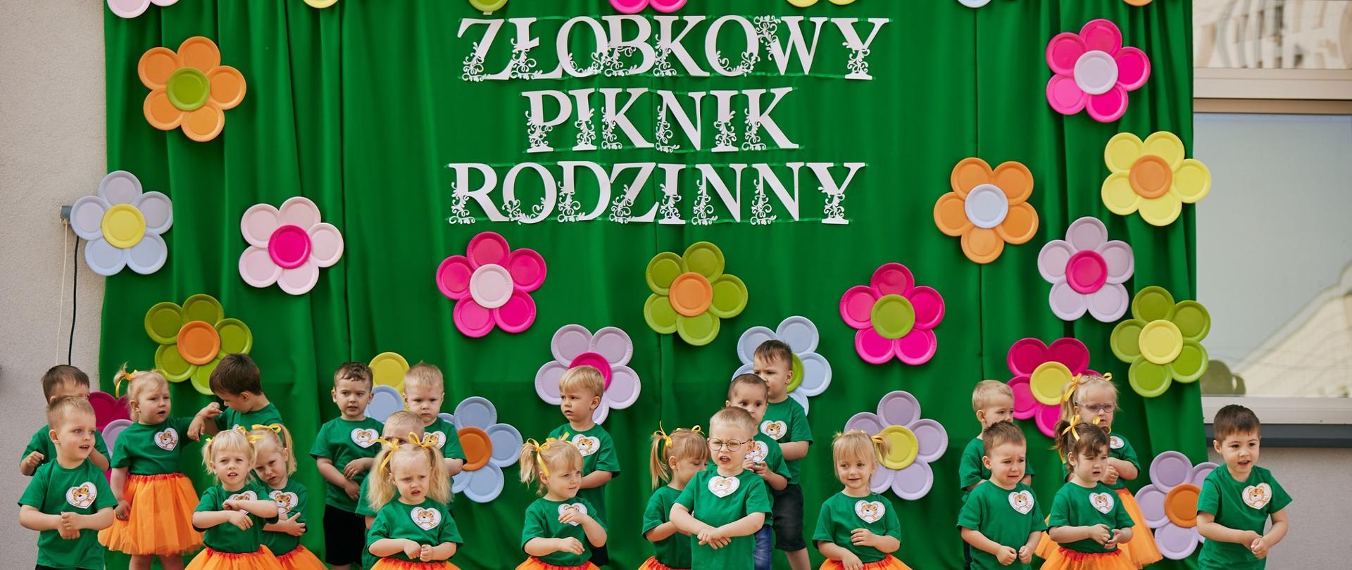 Na zdjęciu widać dzieci z grupy Tygryski. Dzieci prezentują układ muzyczno-taneczny. Wszystkie dzieci stoją na scenie, w tle zielony materiał z kwiatami i napisem Żłobkowy Piknik Rodzinny. Dzieci ubrane są w zielone t-shirty z naklejką z tygryskiem, chłopcy mają czarne spodenki, dziewczynki pomarańczowe spódniczki