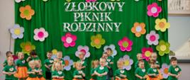 Na zdjęciu widać dzieci z grupy Tygryski. Dzieci prezentują układ muzyczno-taneczny. Wszystkie dzieci stoją na scenie, w tle zielony materiał z kwiatami i napisem Żłobkowy Piknik Rodzinny. Dzieci ubrane są w zielone t-shirty z naklejką z tygryskiem, chłopcy mają czarne spodenki, dziewczynki pomarańczowe spódniczki