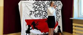 Uczennica śpiewa do mikrofonu pieśń patriotyczną, w tle dekoracja: na białym płótnie biało-czerwona mapa polski, na niej czarna sylweta drzewa z huśtającą się na huśtawce dziewczynką