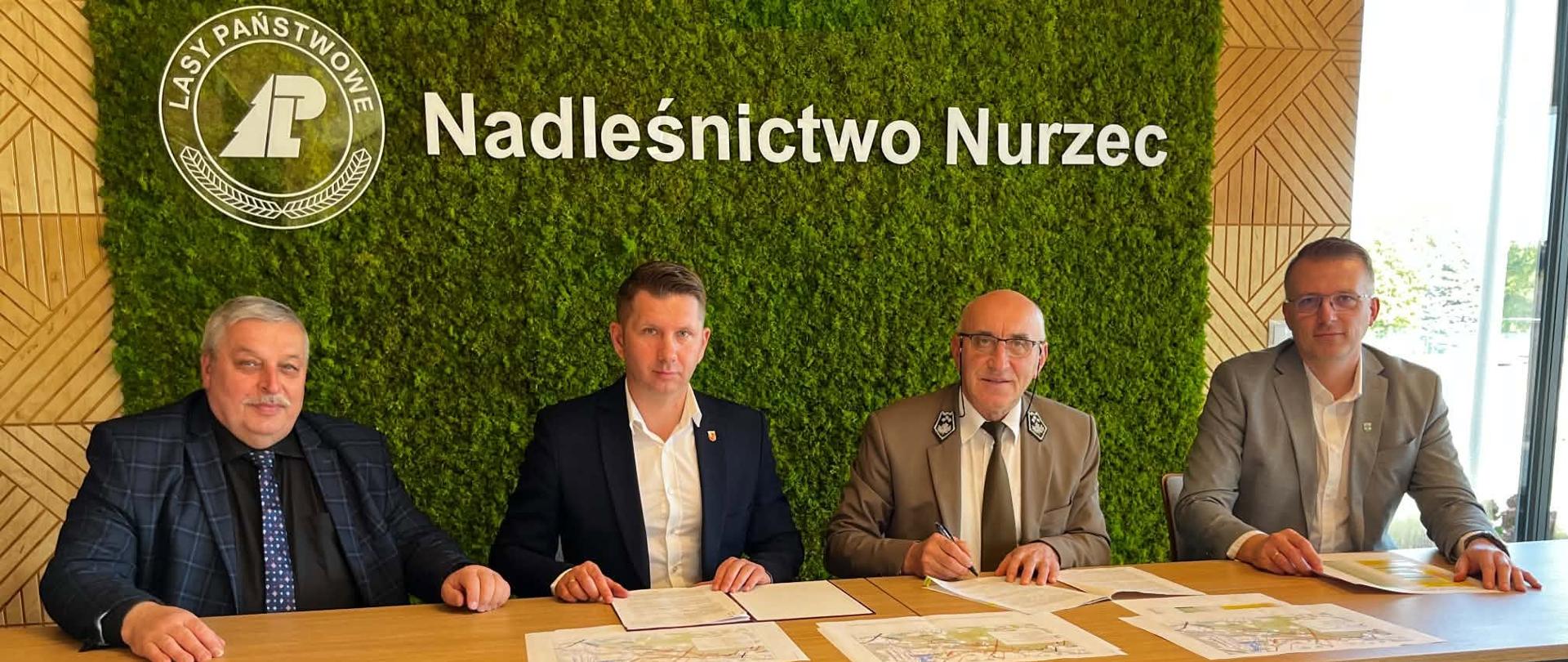 Czterech mężczyzn siedzi przy stole z mapami i dokumentami pod zielonym napisem „Nadleśnictwo Nurzec”.

