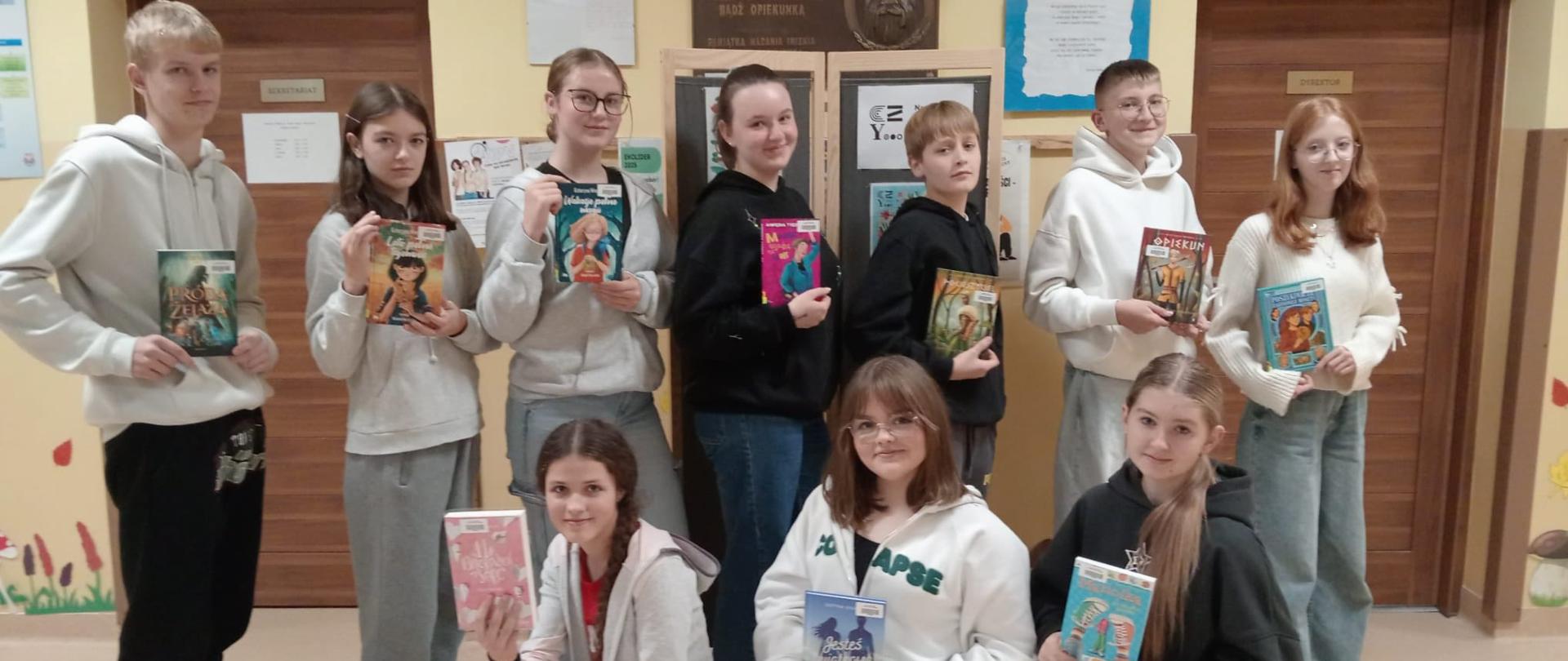 Uczniowie klasy 8 stoją na korytarzu szkolnym pod ścianą upamiętniającą patronkę szkoły Świętą Kingę w rzędzie i trzymają w ręku po jednej książce z nowości zakupionych do biblioteki, przed nimi trzy dziewczynki kucają z książką w ręku